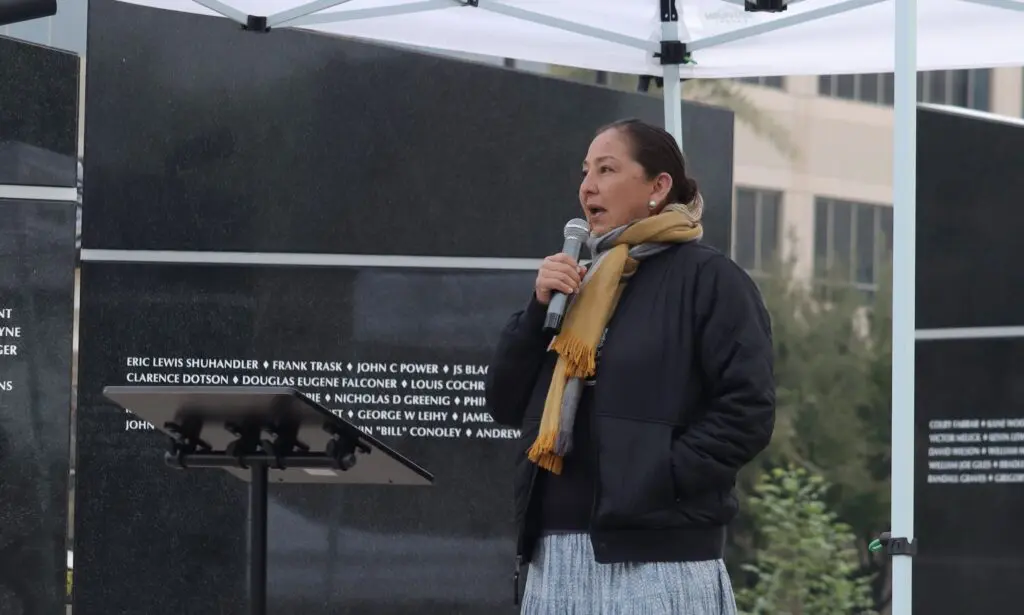 A woman speaks into a microphone at an outdoor event, standing before a large memorial wall with engraved names.