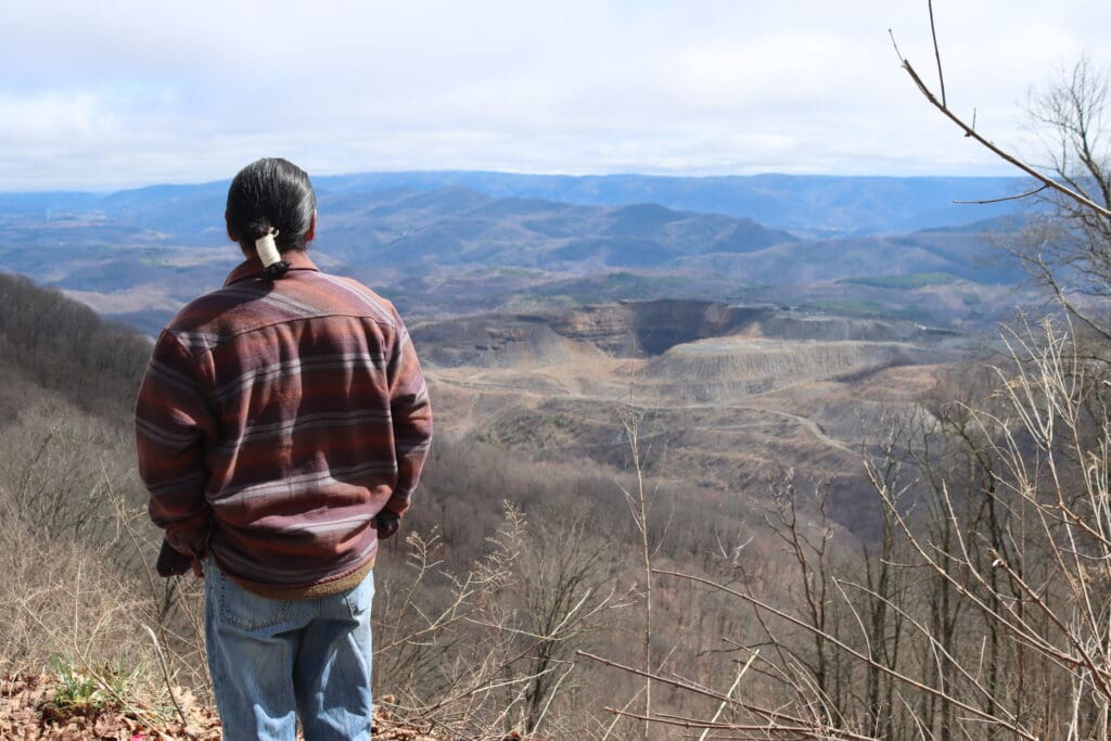 A person in a plaid jacket stands at a scenic overlook enjoying a vast view of rolling hills and bare trees under a cloudy sky.